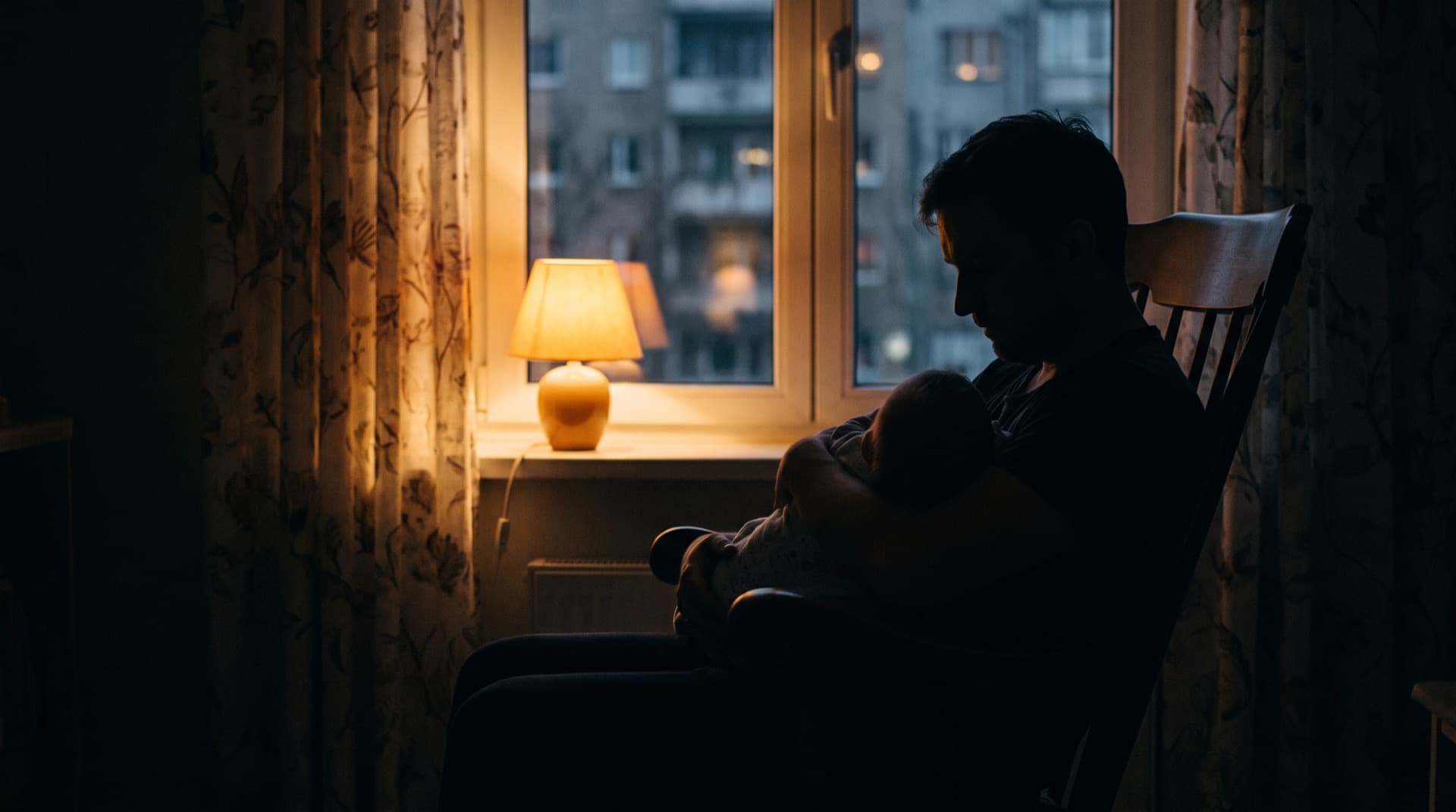 A parent rocks a child by a lamp-lit window, late at night.